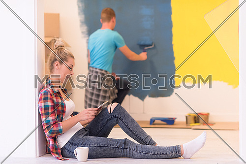 Happy couple doing home renovations, the man is painting the room and the woman is relaxing on the floor and connecting with a tablet