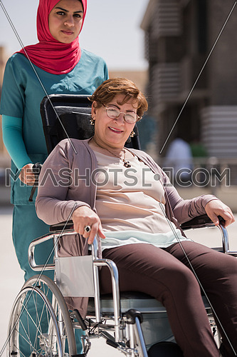 older sick woman in wheelchair with young middle eastern  nurse in front of a large modern hospital