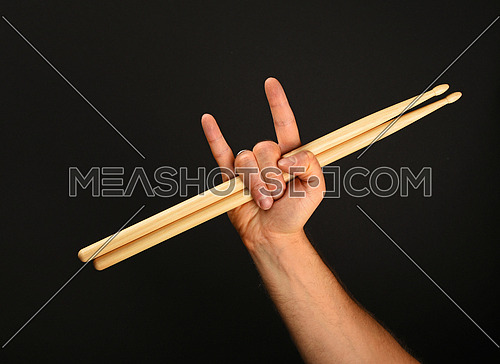 Man hand holding two wooden drumsticks with devil horns rock metal gesture sign over black background, front view