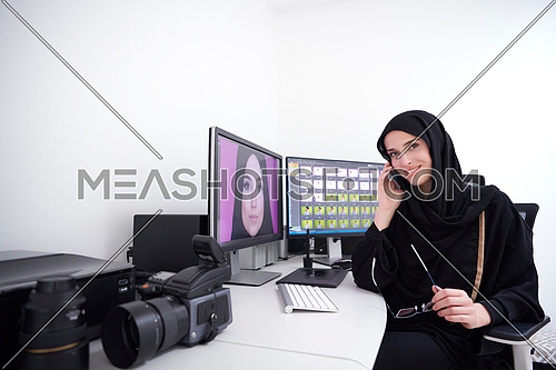 Muslim female graphic designer talking on the phone. Freelancer editing photos on two monitors. Hijab girl on the work place