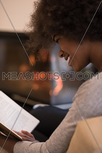 Young beautiful african american woman sitting in front of fireplace at home on a cold autumn day and reading book