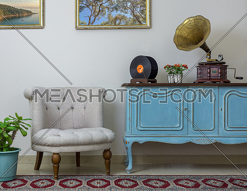 Vintage interior of retro off white armchair, vintage wooden light blue sideboard, old phonograph (gramophone) and vinyl records on background of beige wall, tiled porcelain floor, and red carpet