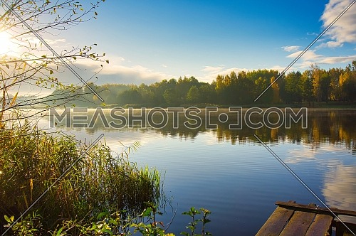 Sunset over a tranquil rural lake with reflections on the water of the surrounding trees and forests with a rustic wooden jetty in the foreground and mist in a scenic landscape