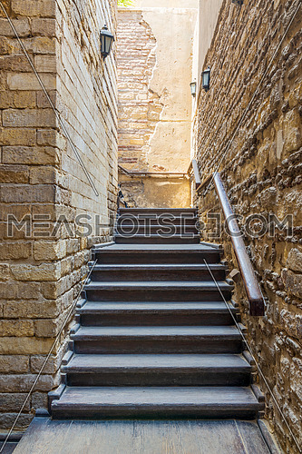 Architectural detail of outdoor wooden old weathered narrow staircase going up with wooden handrail, between two stone bricks walls, in abandoned building