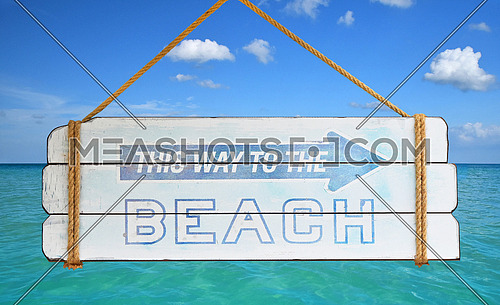 Close up old wooden directional arrow sign with this way to the beach text hanging over background of blue sky and sea