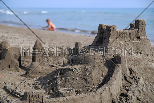 A sand castle in foreground  while a baby playing by the beach