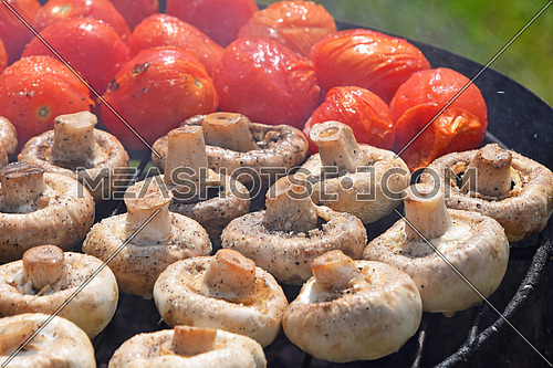 Vegetables in salt and spices being cooked on round char grill outside, white champignons portobello mushrooms and red small tomatoes