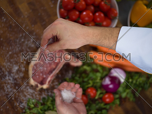 Master Chef hands putting salt on juicy slice of raw steak with vegetables around on a wooden table