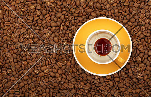 Empty black coffee yellow cup with saucer on background of roasted coffee beans, elevated top view, close up
