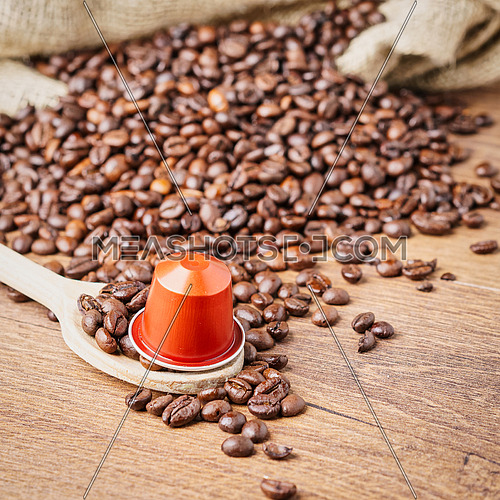 In the foreground a coffee capsule on wooden spoon and  roasted coffee beans with burlap sack on blur wooden background,top view.