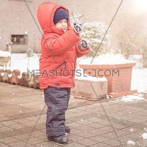 Infant boy smiles cheerfully while it's snowing,dressed in a red winter jacket and a woolen hat.