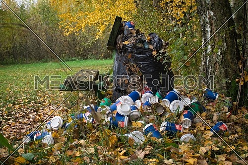 Pile of disposable coffee cups alongside a rustic wooden bench in a dense stand of deciduous forest in autumn in an environmental pollution concept