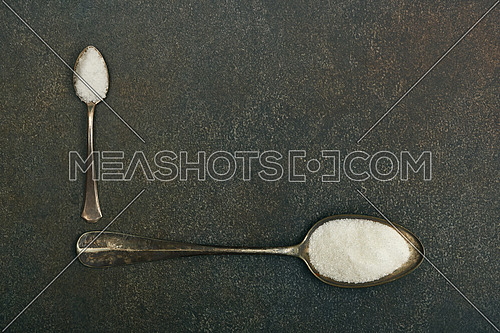 Close up flat lay two vintage metal spoons of white sugar and sea salt on dark grunge stone table surface with copy space, elevated top view, directly above