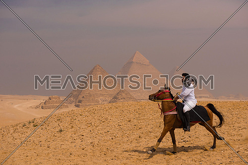young  Egyptian   riding arabian horse in desert,  giza platou with grand pyramids in background