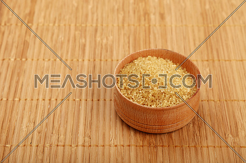 Small wooden round bowl full of brown cane sugar on bamboo mat background