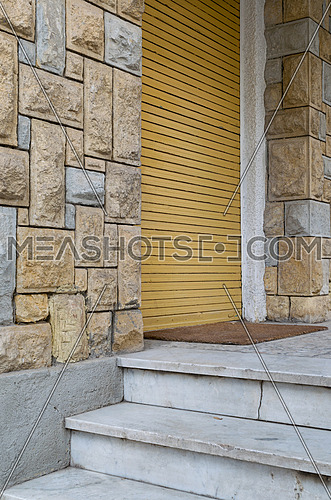 Angled view of stone bricks colored wall, three white marble steps, and closed yellow vertical roller door