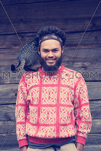 portrait of young hipster man with beard and cat in front of wooden house