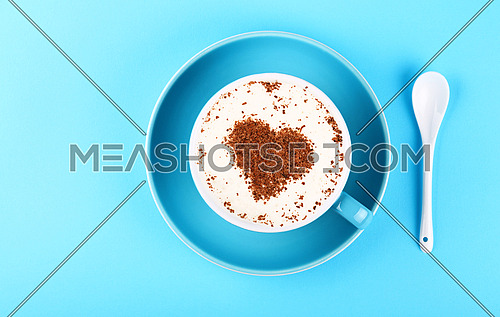 Close up one full cup of latte cappuccino frothy coffee with heart shaped chocolate and saucer with white spoon over pastel blue paper background, elevated top view, directly above