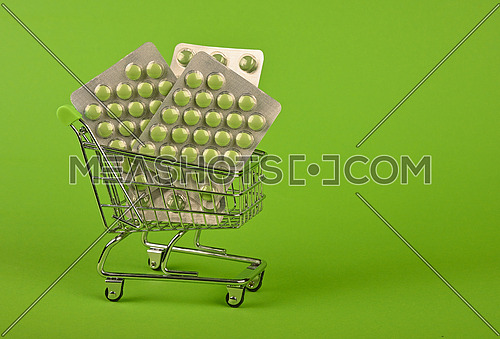 Close up several different blister packs of pills in small shopping cart over green background, concept of online medicine order delivery, low angle view