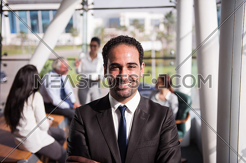Portrait of a male executive wearing eye glasses and a group meeting is taking place at the background in a bright office
