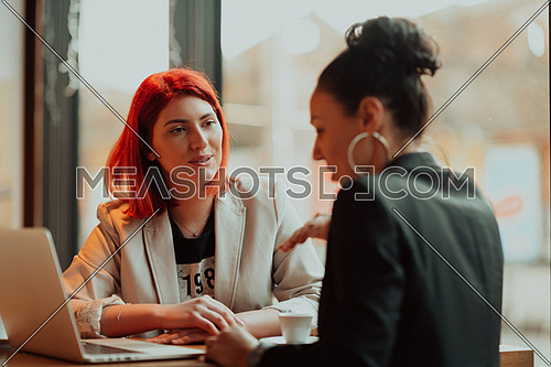 One-on-one meeting.Two young business women sitting at table in cafe. Girl shows colleague information on laptop screen. Girl using smartphone, blogging. Teamwork, business meeting. Freelancers working.