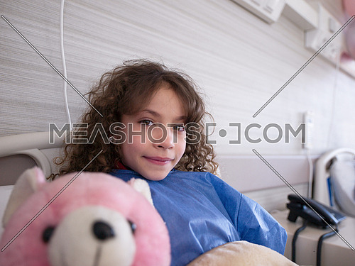 young girl lies in a hospital bed while hugs pink teddy bear