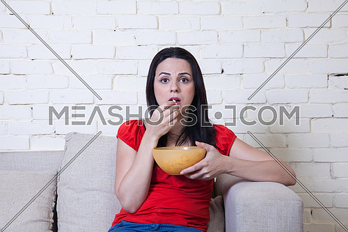 A young woman sitting on a coach eating popcorn shocked