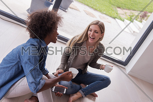 Two young laughing multiethnic women sit on the floor near the window and enjoy while drinking coffee using smartphone in luxury home