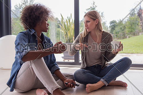 Two young laughing multiethnic women sit on the floor near the window and enjoy while drinking coffee using smartphone in luxury home