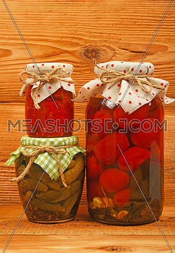 Three glass jars of homemade conserves, eggplant pepper salad, pickled tomatoes and cucumbers with textile decoration over brown unpainted wood
