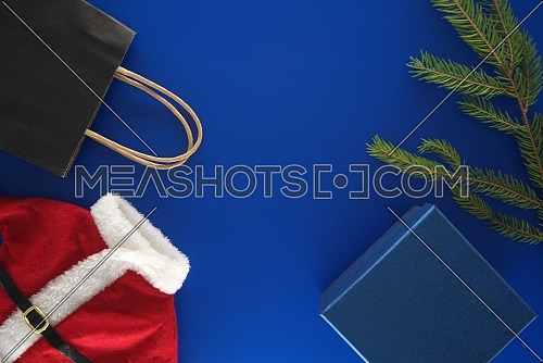 Red Santa Claus suit, black paper shopping bag, gift box and fir branch on blue background with copy space for text