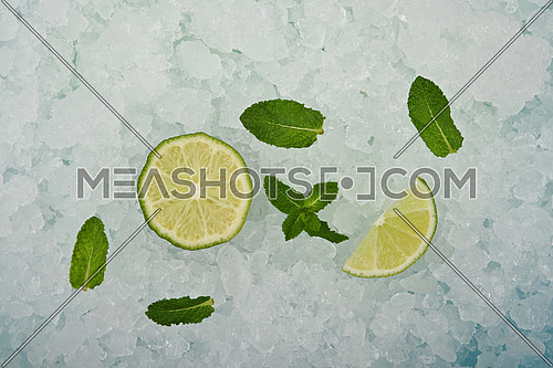 Close up fresh juicy green lime cut half and wedge slice and mint leaves over background of crushed ice, elevated high angle view, directly above