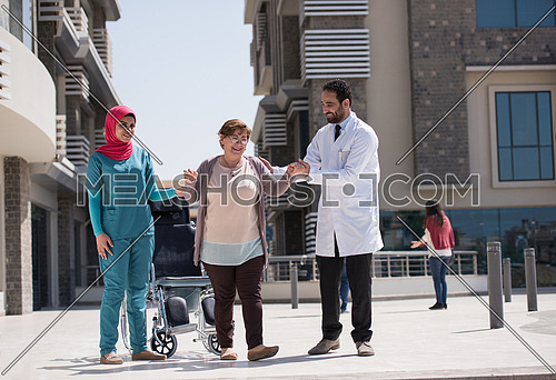elderly sick woman in a wheelchair with medical staff in front of large modern hospital in the Middle East