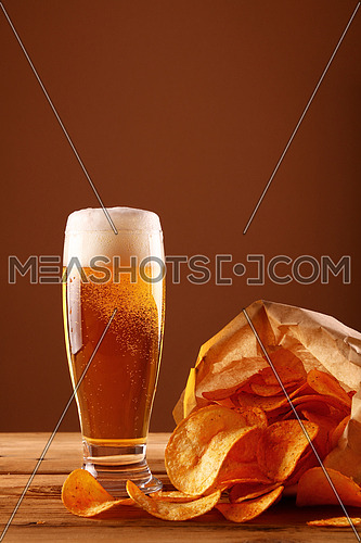 Close up one glass of lager beer with white froth and bubbles and paper bag of potato chips on wooden table over dark brown background with copy space, low angle side view