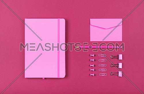 Neatly organized stationery flat lay of pastel pink notebook, envelope, pencil, office clips, binders and pins in order over dark purple pink background, elevated top view, directly above