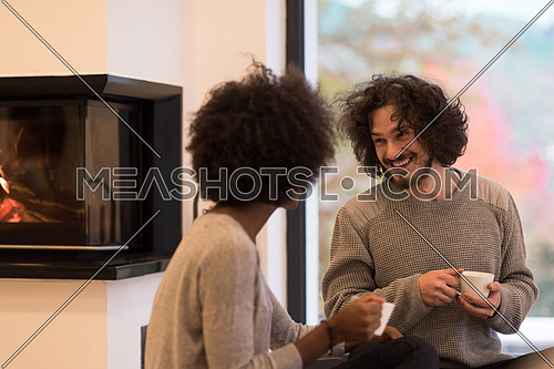 Young romantic multiethnic couple sitting on the floor in front of fireplace at home, looking at each other, talking and drinking coffee autumn day