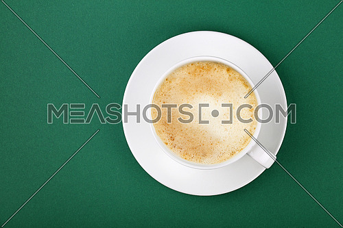 Close up one full white cup of frothy latte cappuccino coffee on saucer on green table background, elevated top view, directly above
