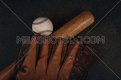 Close up one old baseball ball, wooden bate and worn leather vintage glove on grunge dark brown background, elevated top view, directly above