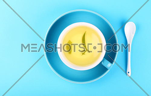 Close up one full cup of green oolong or herbal tea on saucer with white spoon over pastel blue paper background, elevated top view, directly above