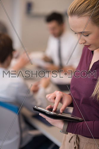 Blonde businesswoman in casual  clothes working on tablet computer at modern startup business office interior. Young people group on team meeting blured  in background