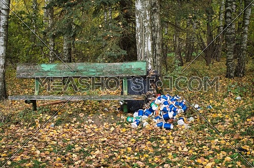 Pile of cans and containers alongside a rustic wooden bench in a dense stand of deciduous forest in autumn in an environmental pollution concept