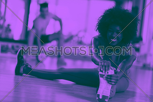 happy young african american woman in a gym stretching and warming up before workout young mab exercising with dumbbells in background duo tone filter