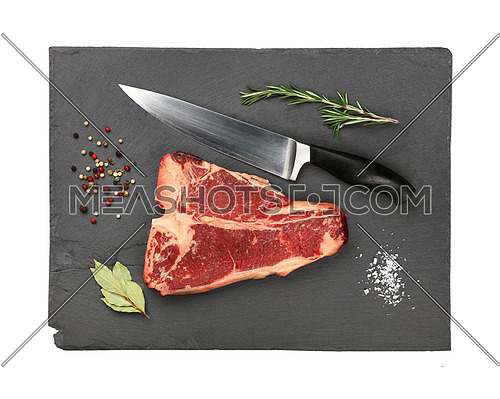 Close up one marbled raw porterhouse T-bone beef steak with rib bone on black slate cutting board with knife and spices to season, isolated on white background, elevated top view, directly above