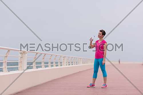 Fitness woman drinking water after running at the promenade by the sea. Thirsty sport runner resting taking a break with water bottle drink outside after training. Beautiful fit sporty girl.