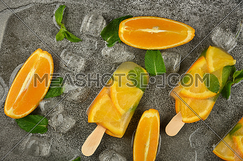Close up three fruit ice cream popsicles with fresh orange slices, green mint leaves and ice cubes on gray table surface, elevated top view, directly above