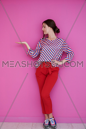 Portrait of happy smiling young beautiful woman in a presenting gesture with open palm isolated on pink background. Female model in modern fashionable clothes posing in the studio