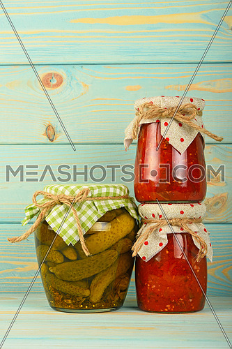 Two glass jars of homemade eggplant pepper salad and pickled cucumbers with green checkered textile decoration over blue painted wooden surface