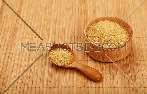 Wooden round scoop spoon and small bowl of brown cane sugar on bamboo mat background