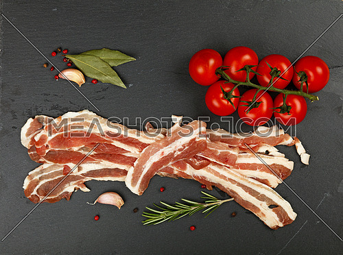 Raw pork bacon slices, rashers, spices, peppercorns, garlic, bay laurel leaves and red fresh cherry tomatoes on black slate board, close up, elevated top view