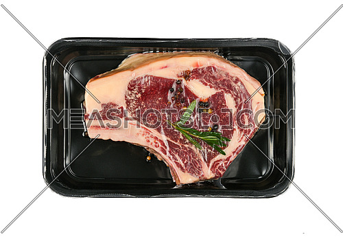 Close up one marbled raw ribeye beef steak with rib bone vacuum sealed with black plastic film, isolated on white background, elevated top view, directly above